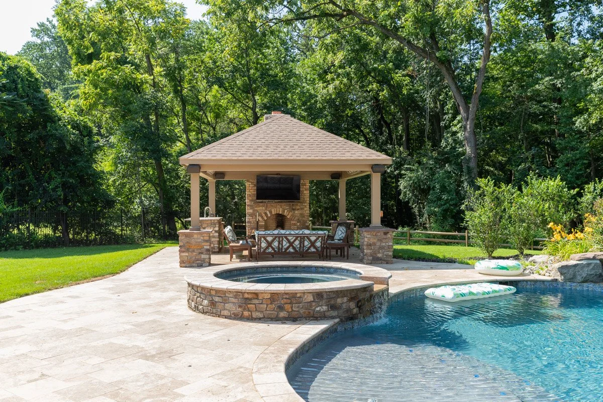 Backyard with a swimming pool, hot tub, and a poolside pavilion with outdoor furniture and a TV, surrounded by green trees and garden plants.
