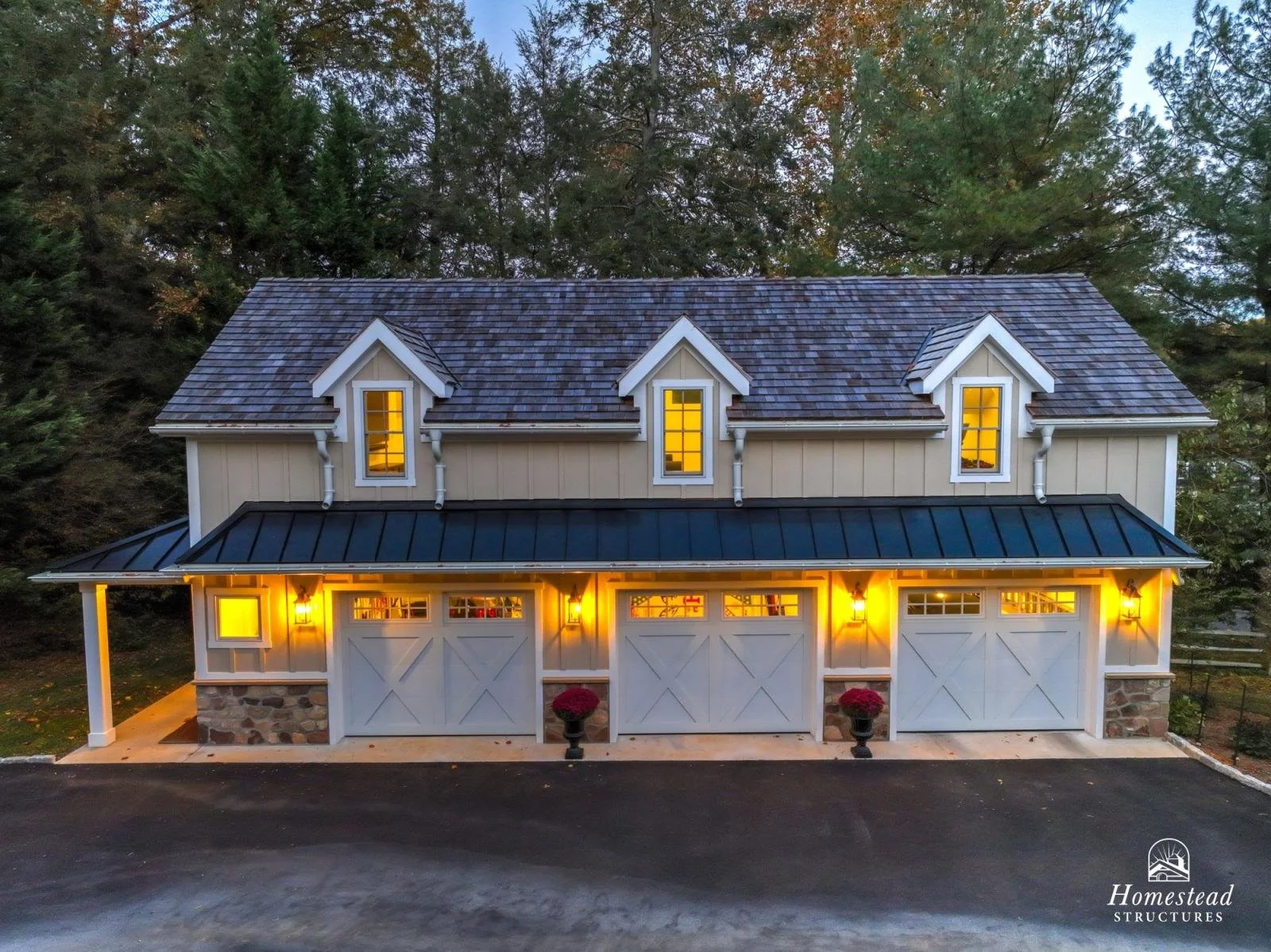 A two-story garage building with three garage doors, exterior lights, a peaked roof with dormer windows, surrounded by trees.