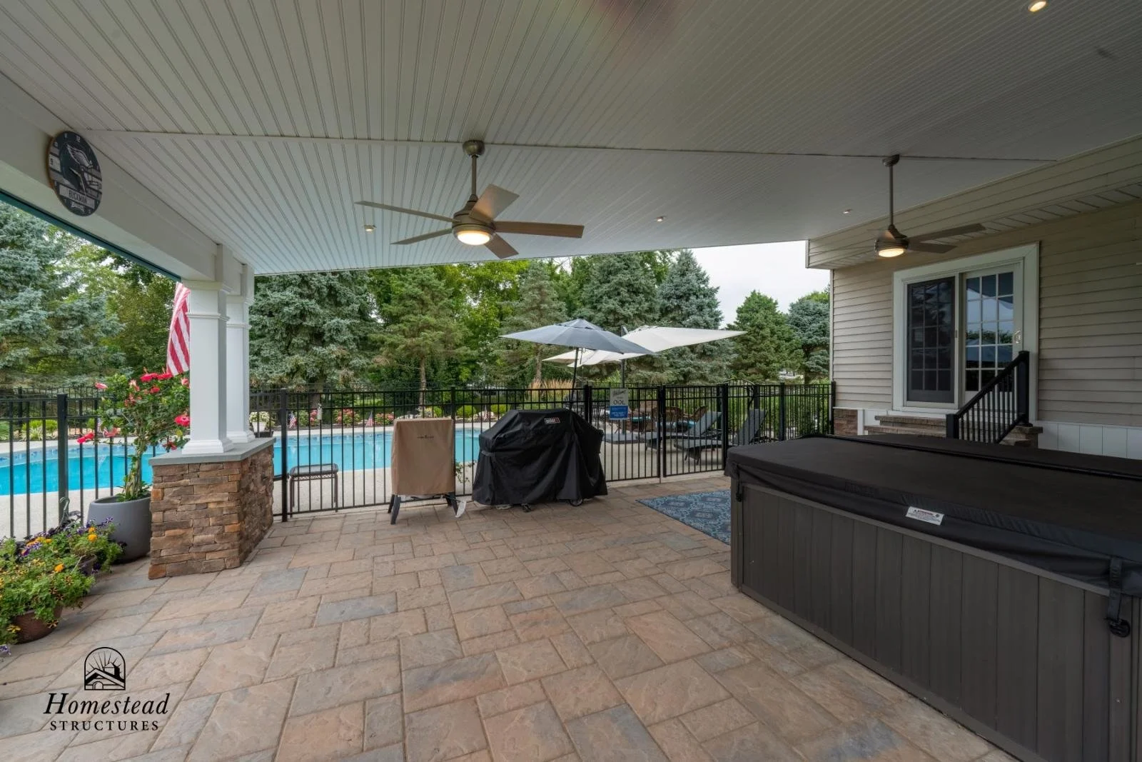 Covered outdoor patio area with ceiling fans, view of a swimming pool, umbrellas, lounge chairs, and surrounding trees.