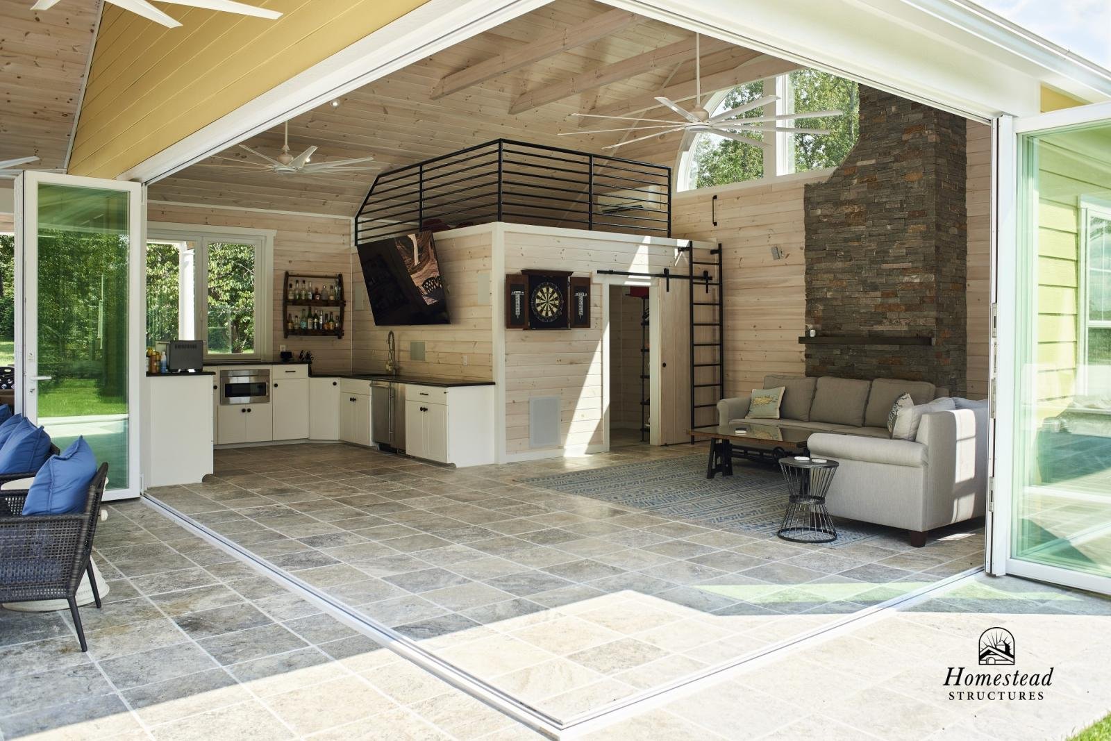 Open-concept living space with kitchen and seating area featuring a large stone fireplace, a sectional sofa, dartboard on the wall, and skylights and large windows letting in natural light. Tile flooring and wooden ceiling with fans.