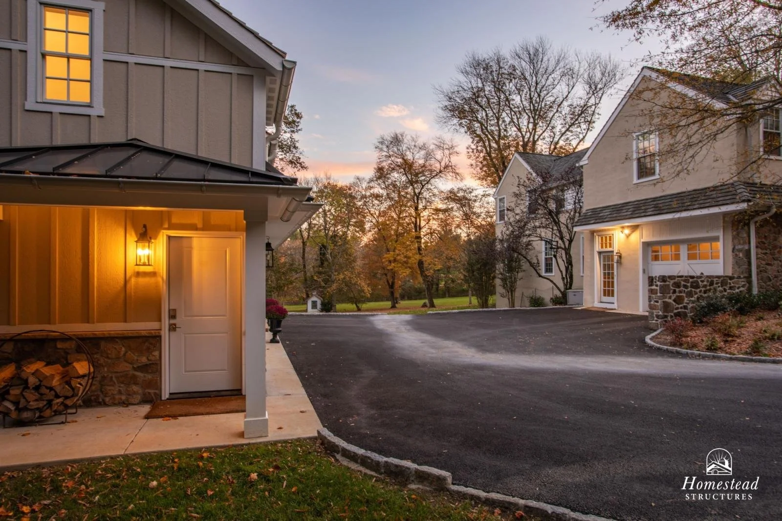 Photo of a residential neighborhood at sunset featuring houses with lit outdoor lights, a driveway, trees with fall foliage, and a small white shed in the background.