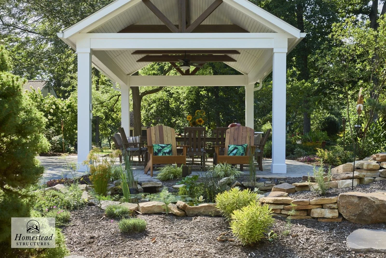 A backyard with a white wooden pavilion covering a seating area with wooden chairs and a table, surrounded by lush greenery and landscaped garden beds with rocks, plants, and flowers.