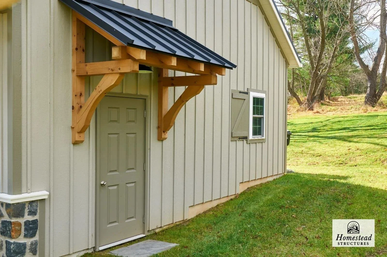 Exterior view of a building with beige vertical siding, a gray door, a small window with gray shutters, and a wooden door canopy with black metal roof supports and shingles, surrounded by grass and trees.