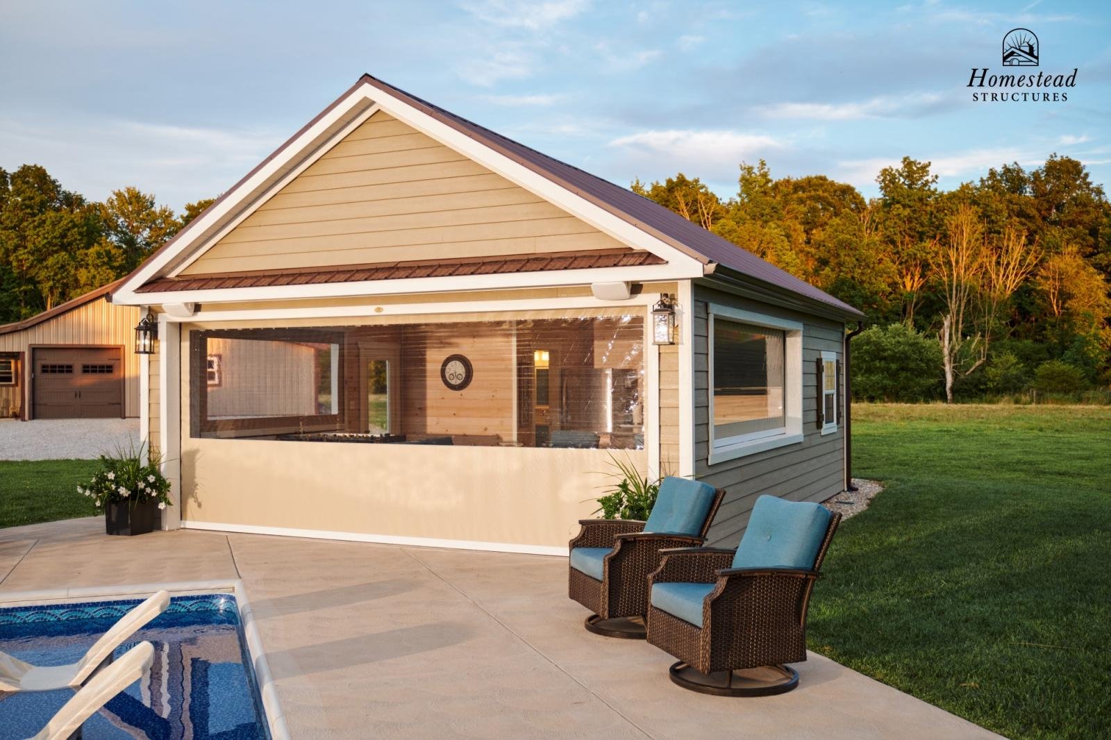 A backyard with a modern house featuring a screened-in porch, two wicker chairs with blue cushions, a swimming pool with white lounge chairs, and a lush green lawn with trees in the background during sunset.