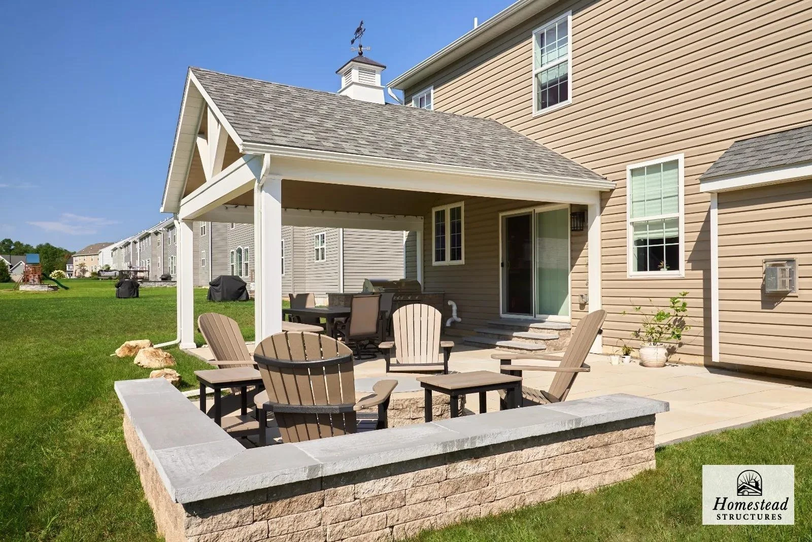 Backyard patio with Adirondack chairs, a dining table with chairs, and a barbecue grill, attached to a beige house with sliding glass door and multiple windows, surrounded by a green lawn and neighboring houses.