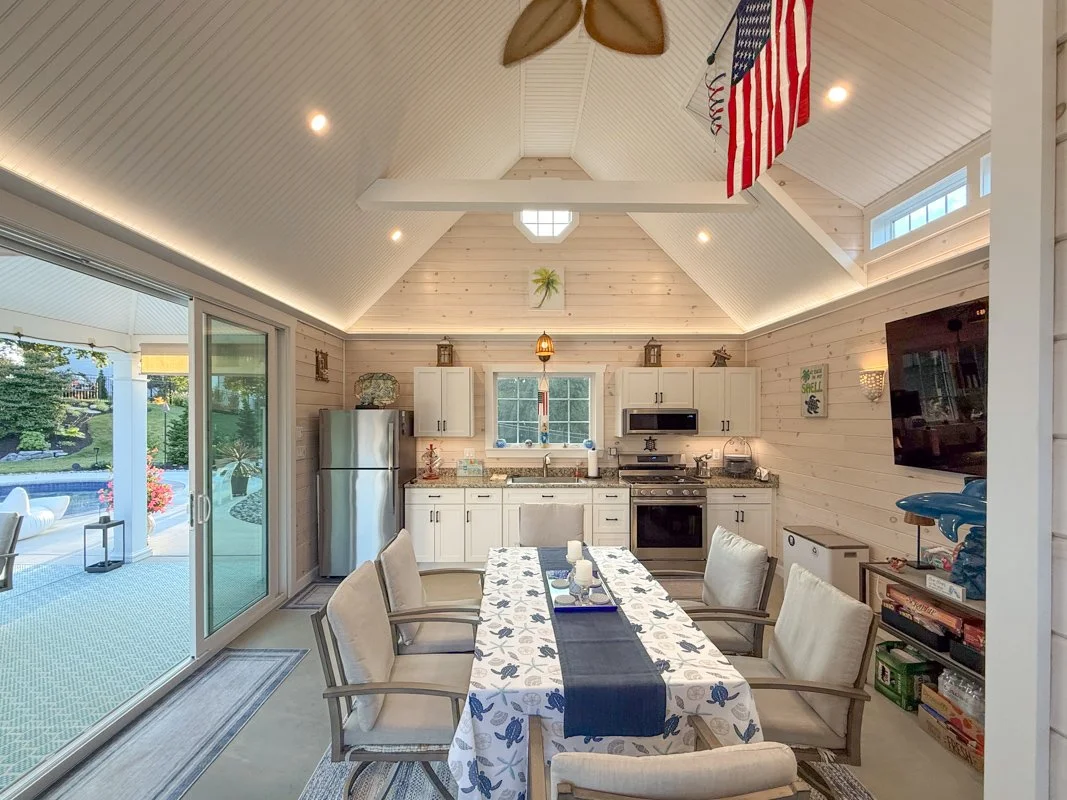 Indoor kitchen and dining area with a light wood-paneled vaulted ceiling, white cabinets, stainless steel refrigerator, stove, and oven. There is a window above the sink, a table with a floral tablecloth and six beige chairs. The room opens to an out