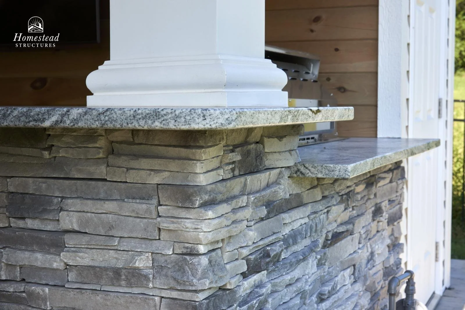 Close-up of stone and granite countertop on a stone and wood outdoor structure with part of a white wooden wall and a pipe underneath
