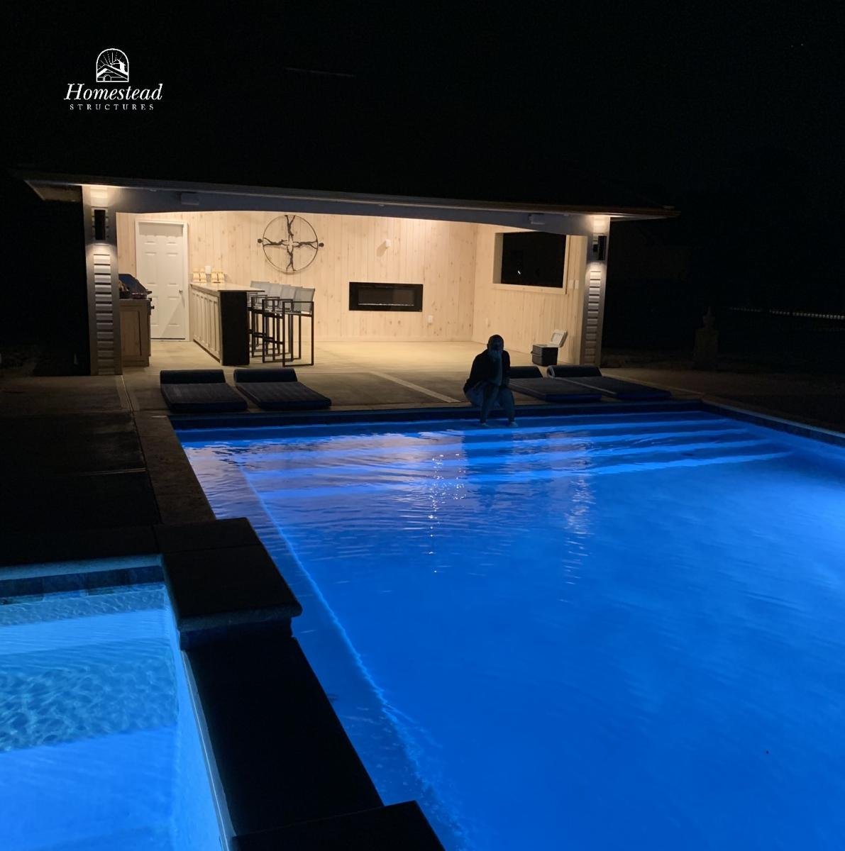 Night view of an outdoor swimming pool illuminated with blue lights, with a person sitting at the edge. In the background, a well-lit outdoor living area with a wooden wall, barstools, and a decorative cross. The scene has a serene and modern ambianc