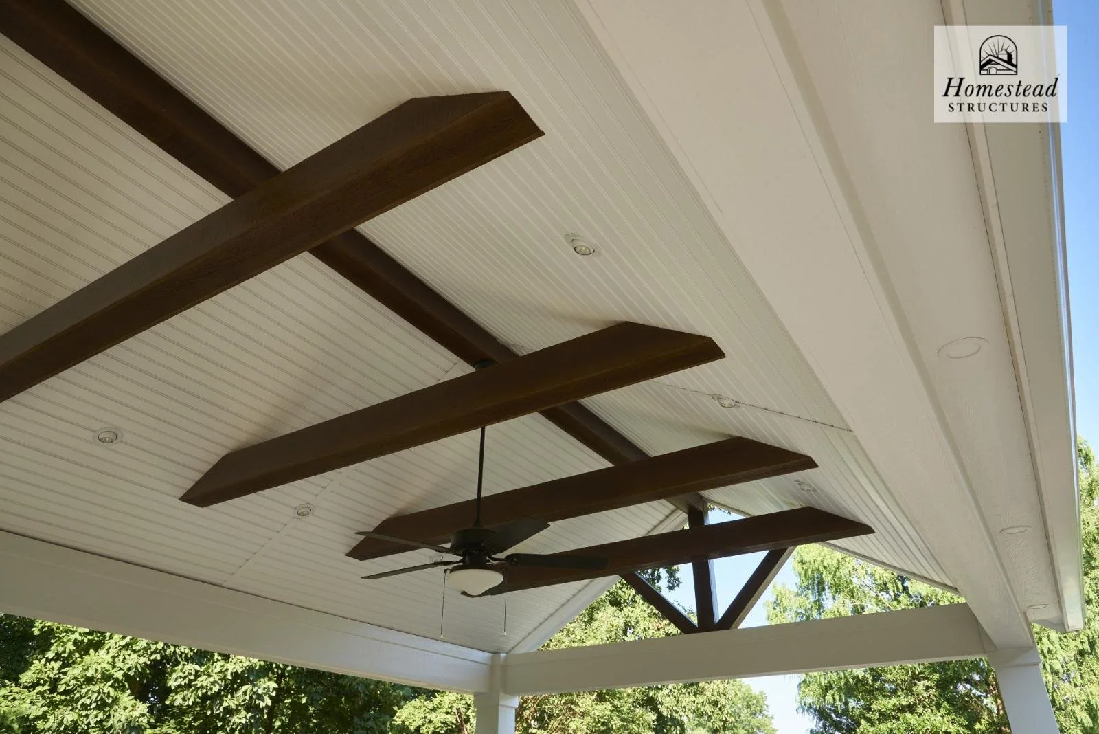 View of a porch ceiling featuring a ceiling fan with four wooden blades, white beadboard ceiling, brown wooden beams, white trim, and visible trees and blue sky in the background.