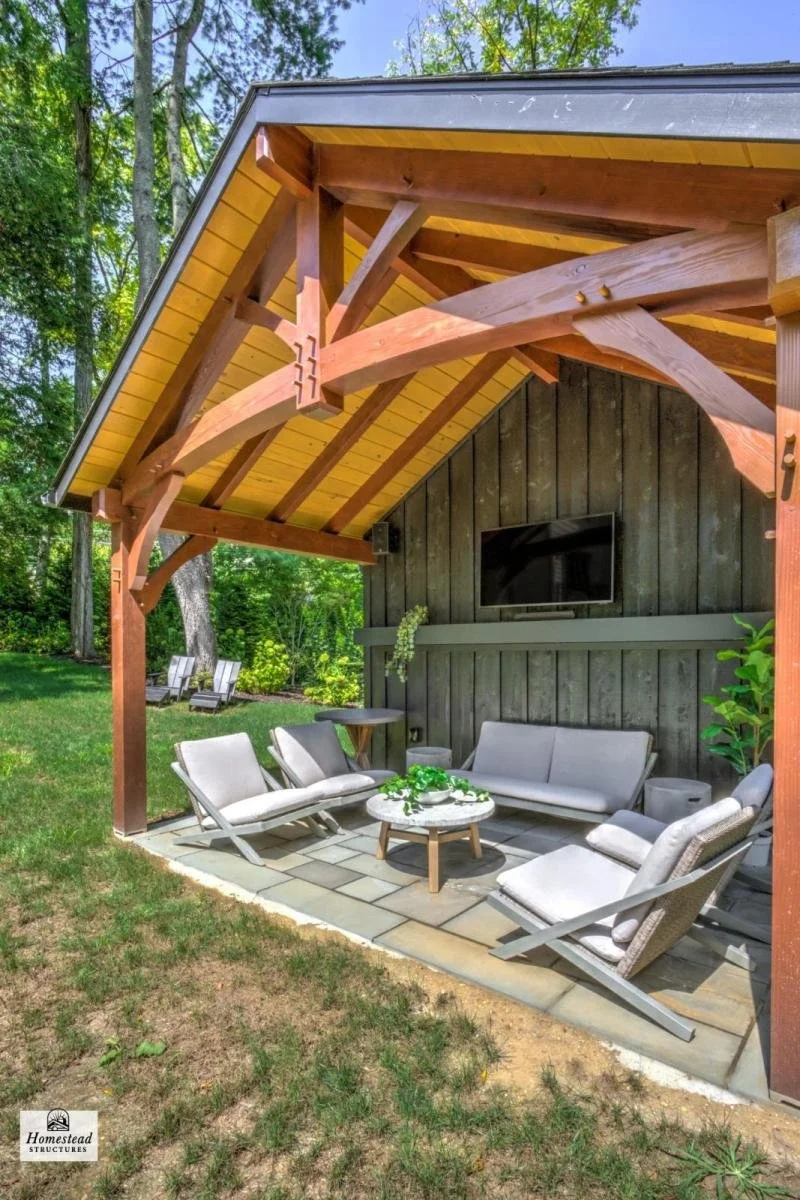 Outdoor patio area with modern furniture under a wooden shelter, surrounded by trees and greenery.