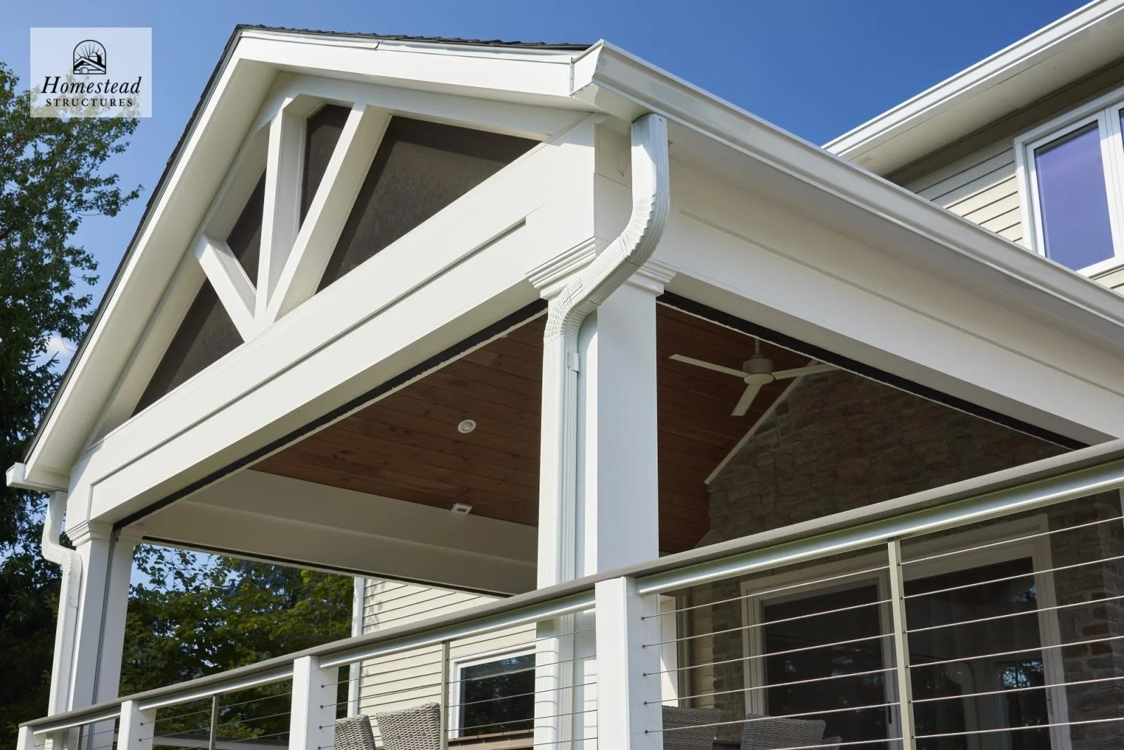 Close-up of a modern two-story house's upper balcony with white railings, a wooden ceiling, and a ceiling fan, with some windows and trees in the background.