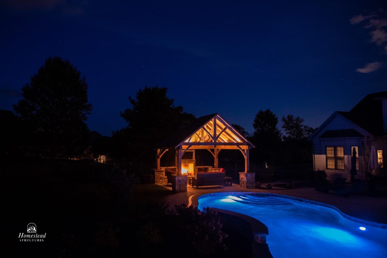Nighttime backyard scene with illuminated pool, stone fire pit, wooden pavilion with a lit fireplace, and surrounding trees.