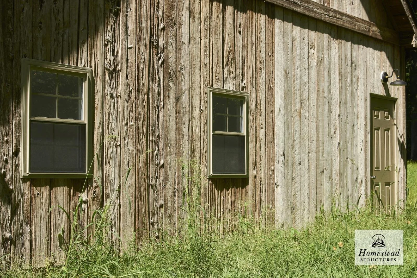 Wooden barn exterior with two small windows and a door, surrounded by grass
