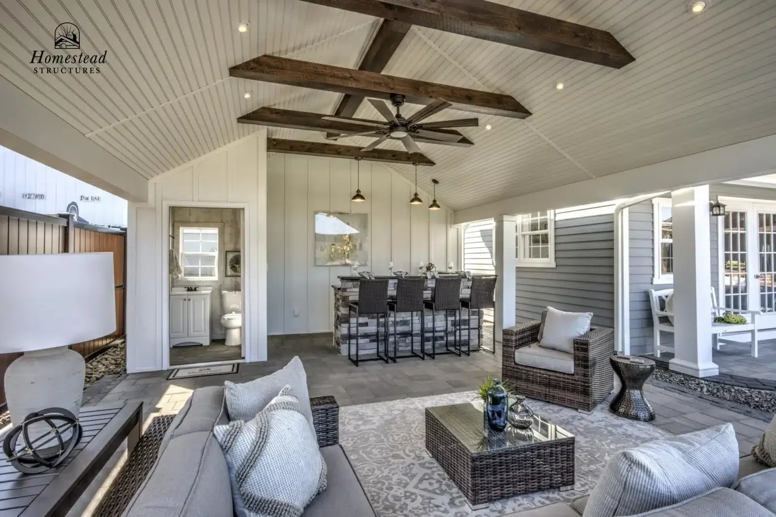 Outdoor covered patio with seating area, bar, and fireplace, featuring wicker furniture, a ceiling fan, and wood accents.