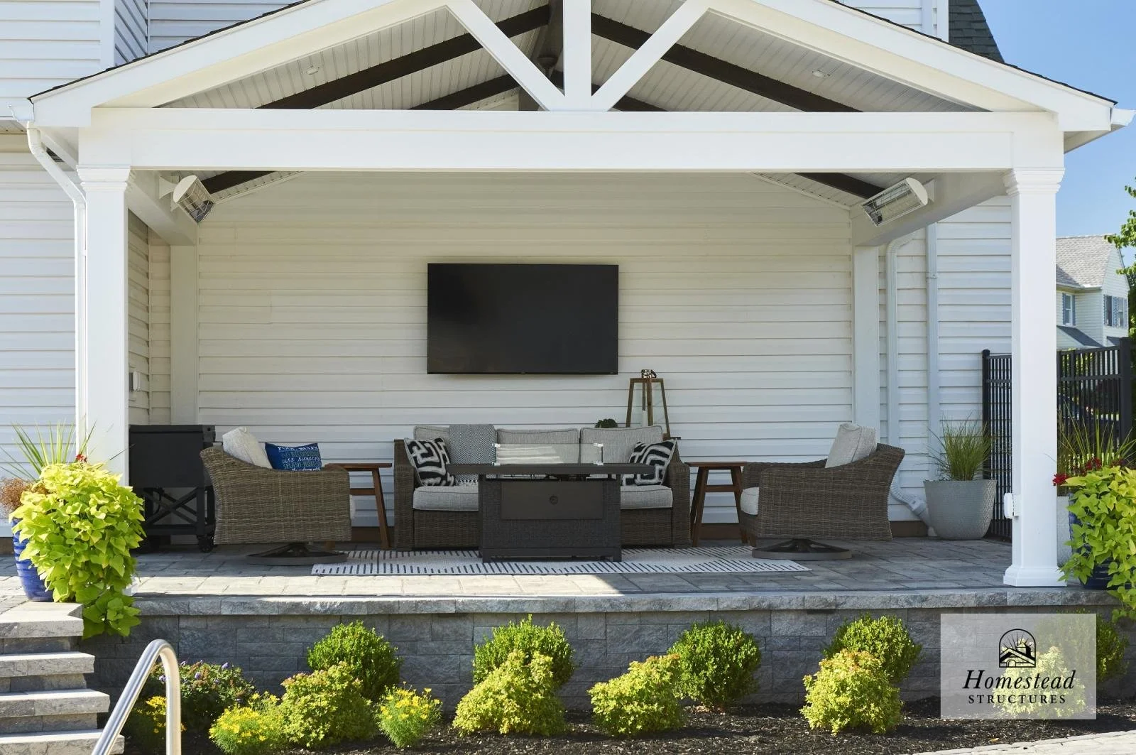 Outdoor patio with a white pavilion, wicker seating with cushions, a wall-mounted TV, and potted plants, surrounded by landscaped bushes.