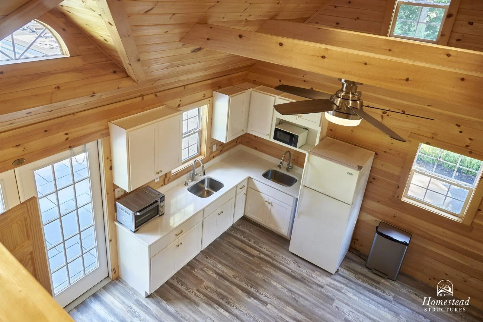 View of a rustic kitchen with natural wood walls and ceiling, white cabinets, two sinks, a refrigerator, microwave, small oven, and a ceiling fan with lights.