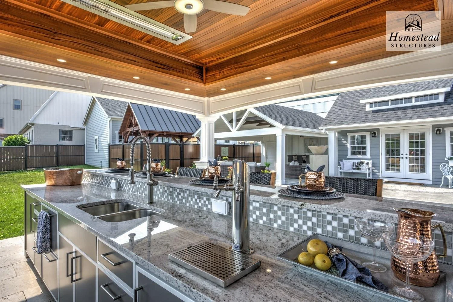 Outdoor kitchen with a granite countertop, sink, and bar seating, overlooking a backyard with a lawn and a yard with small structures and a house with a porch.