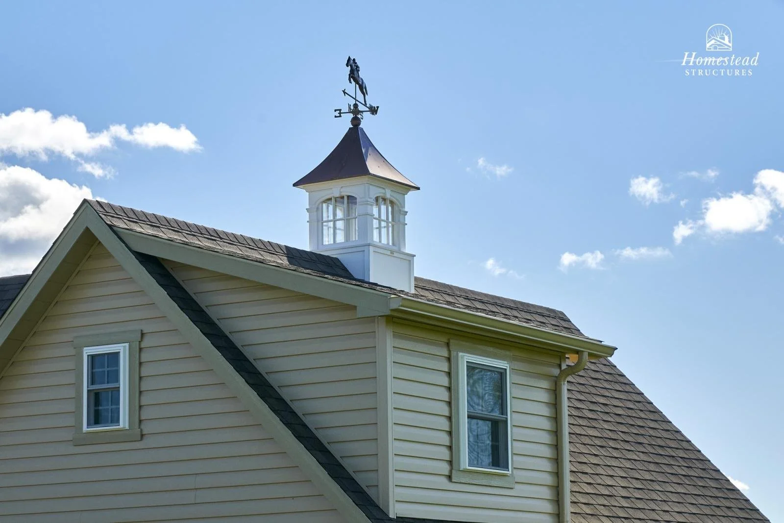 Close-up of a multi-story house with beige siding, white trim, two windows, and a weather vane on the roof against a blue sky with scattered clouds.