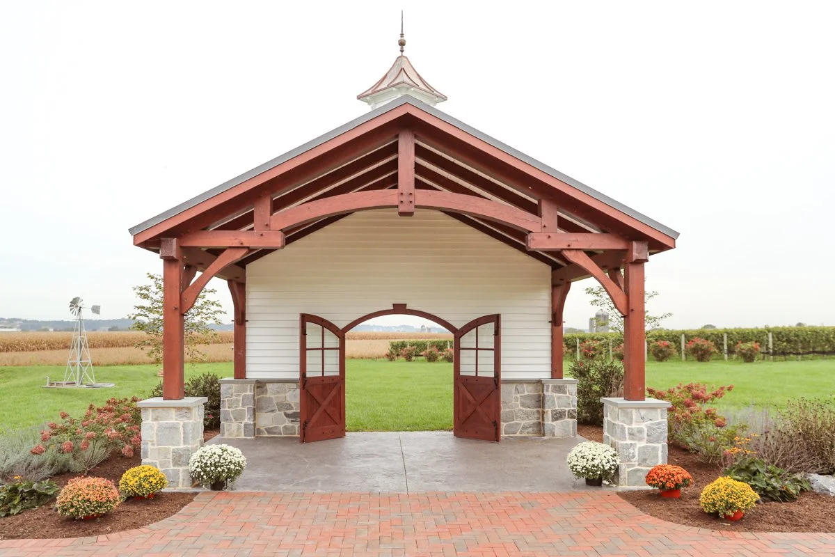 A small barn with a red roof and red wooden doors, stone pillars, surrounded by flower beds and a lawn, with open landscape and a windmill in the background.
