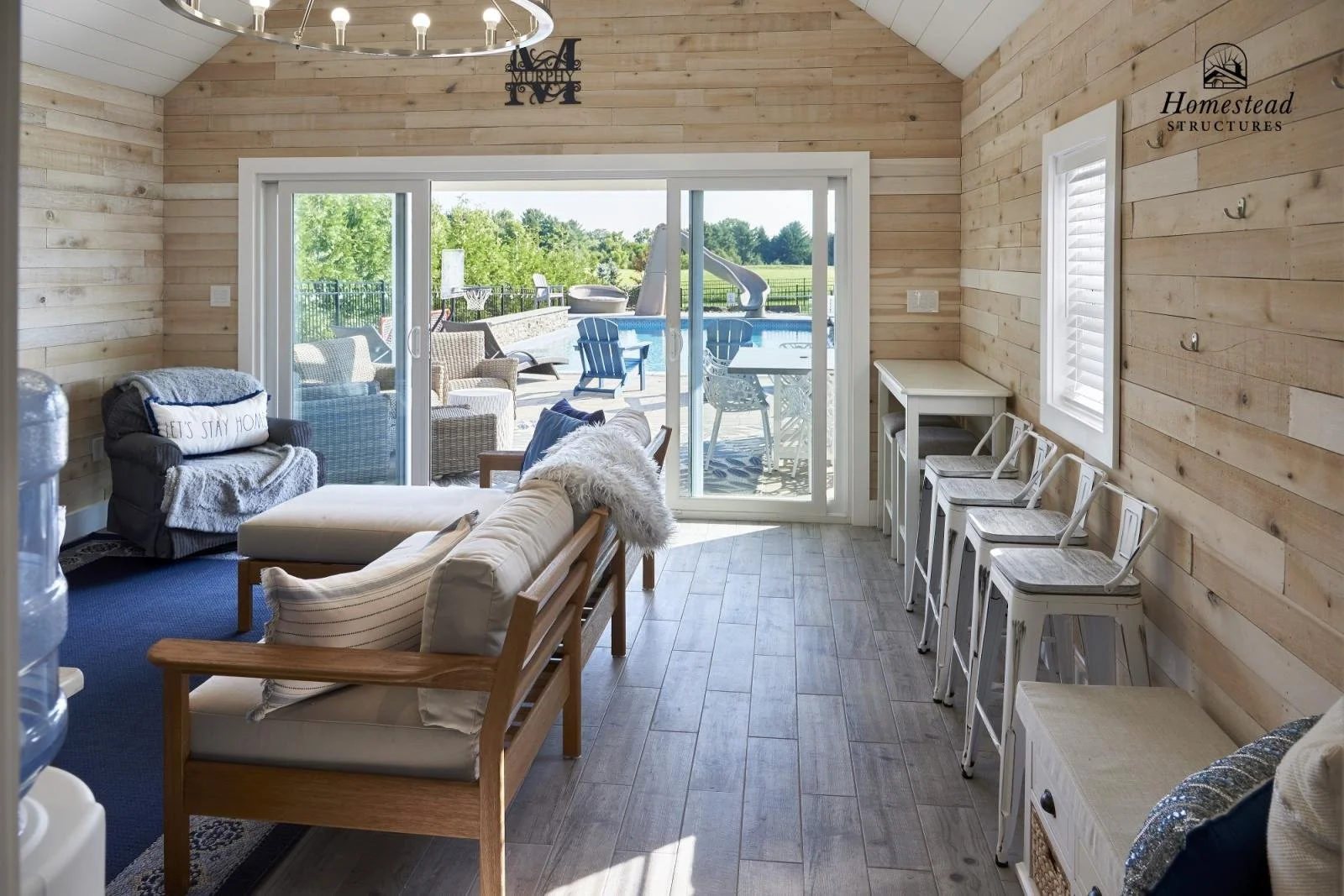 Living room with wooden walls and floor, sliding glass door leading to outdoor patio and pool, and four white bar stools against the wall.