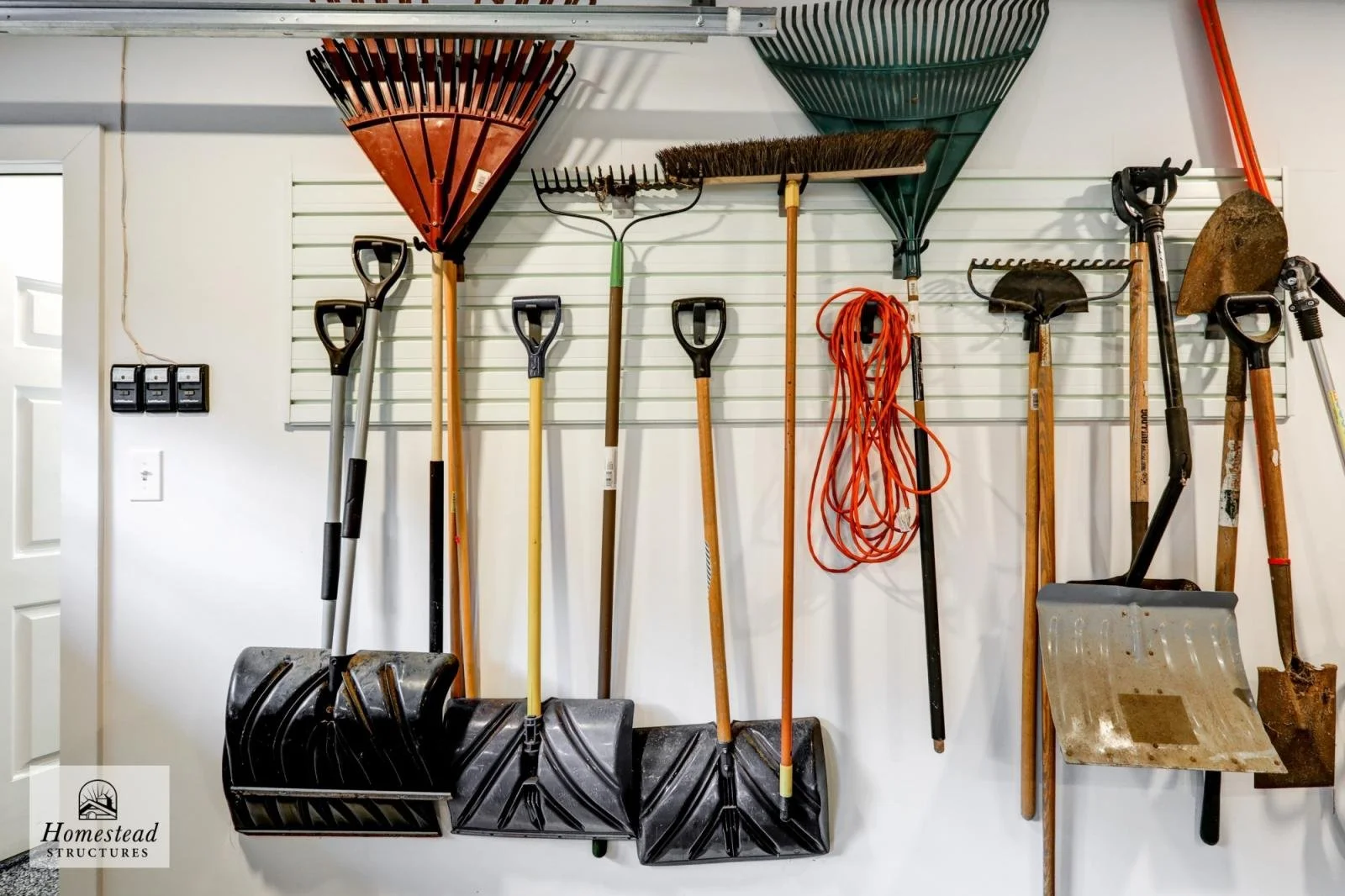 Garden tools hanging on wall, including rakes, shovels, and a snow shovel, along with extension cords on a garage wall.