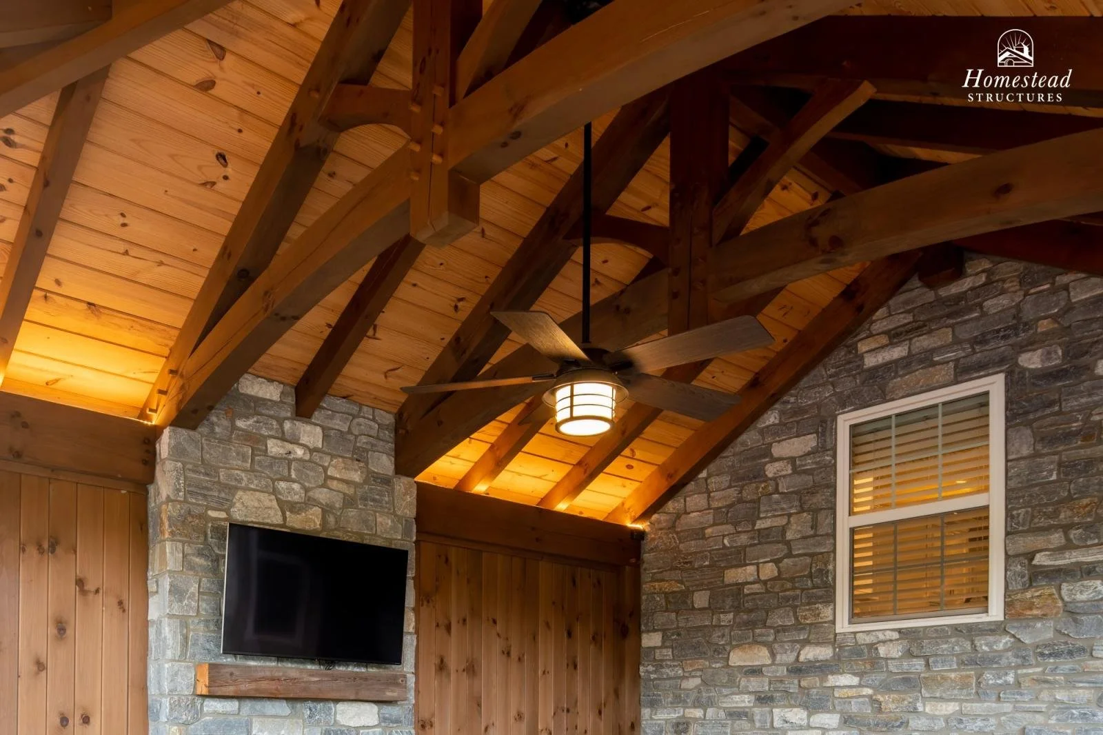 Interior view of a rustic room with a wooden vaulted ceiling, exposed wooden beams, a ceiling fan with lights, a stone accent wall, a flat-screen TV mounted on the wall, and a window with wooden blinds.
