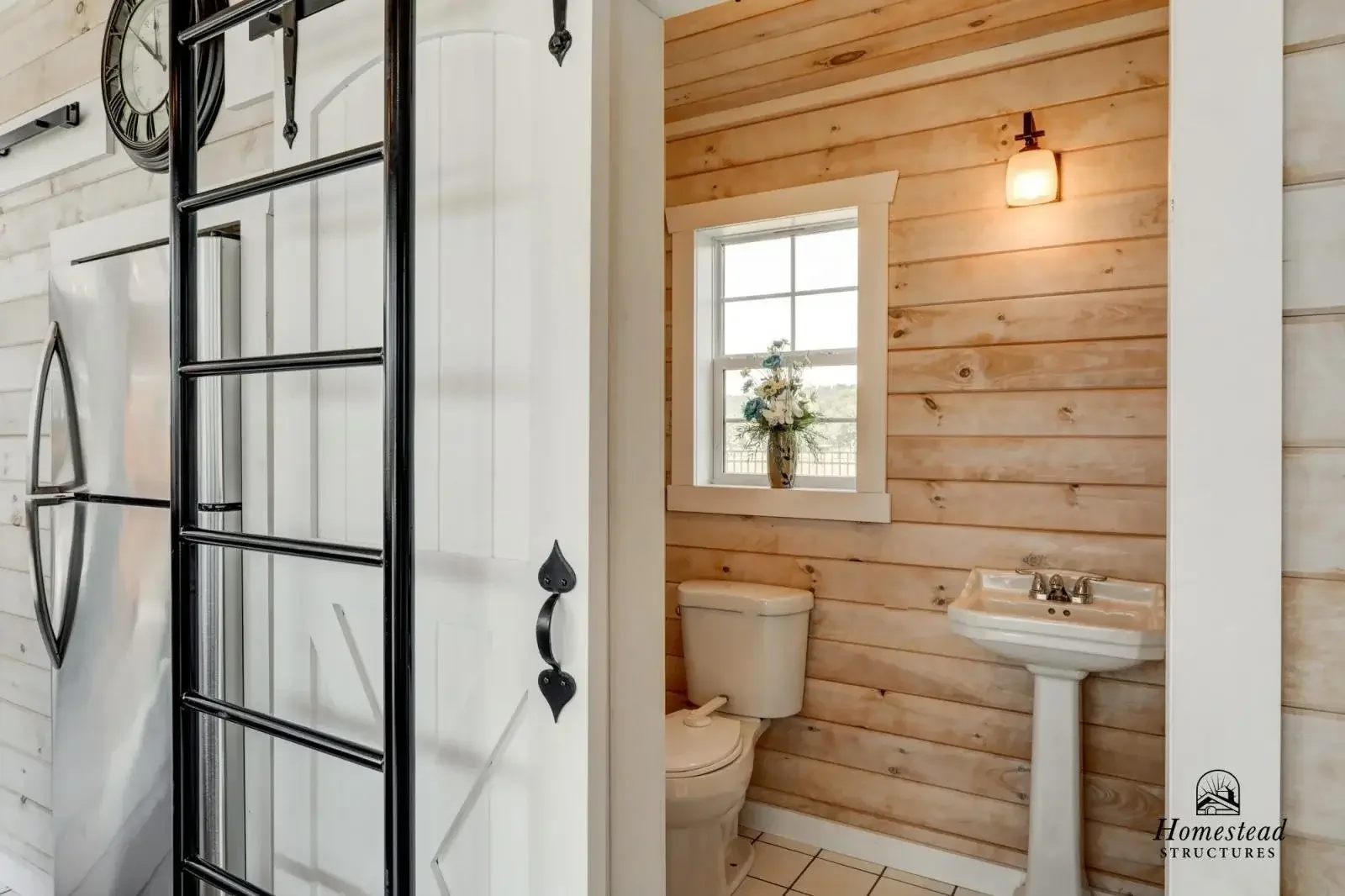 Bathroom with wood-paneled walls, a window with a flower vase on the sill, a toilet, and a small sink. There is a wall-mounted light fixture and a large black metal ladder-style rack on the left.