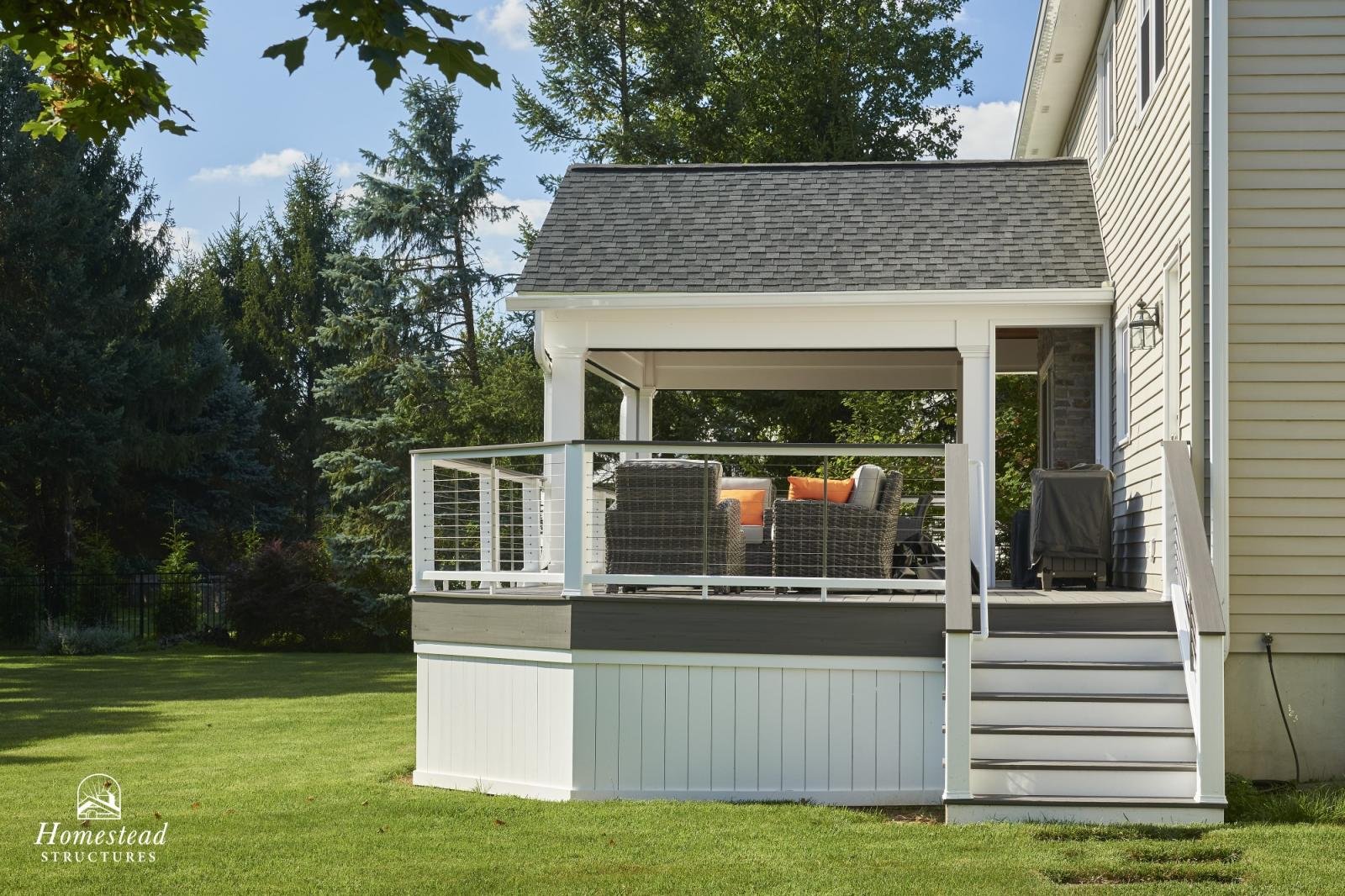 A second-floor porch with wicker furniture and orange cushions, attached to a yellow house with beige siding, overlooking a grassy yard with trees in the background.