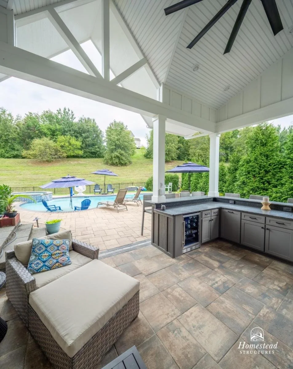 View from a covered patio overlooking a backyard with a swimming pool, lounge chairs with umbrellas, lush green trees, and a grassy hill. Patio area has a woven outdoor sofa with a patterned pillow, a countertop with cabinets, and a ceiling fan.