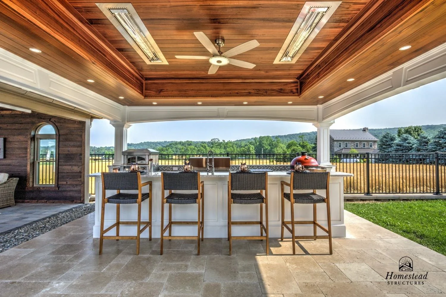 Covered outdoor patio with a white kitchen island, black bar stools, built-in grill, and a view of green fields and trees in the background.