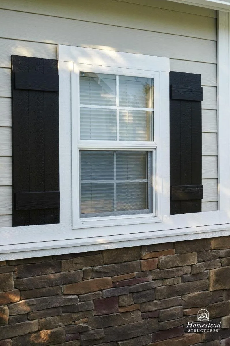 Close-up of a house window with white framing, black decorative shutters on either side, and a stone foundation below.