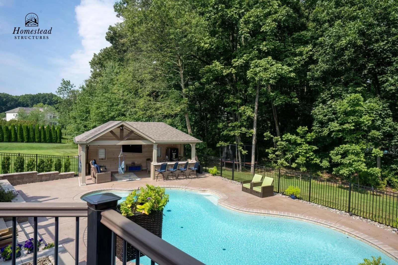 Backyard swimming pool with a poolside patio area, fenced backyard, and dense green trees in the background. There is an outdoor seating area, a bar with four chairs, and a small covered structure with television and outdoor furniture.