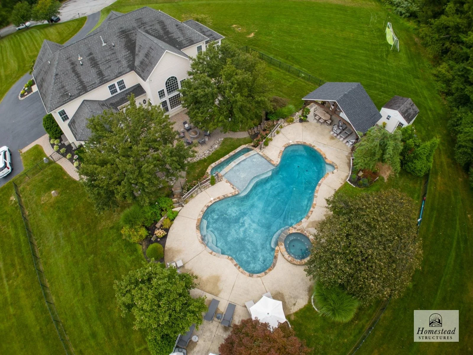 Aerial view of a backyard with a large swimming pool, hot tub, patio furniture, a shaded sitting area, surrounding trees, a grassy lawn, and a two-story house with a dark shingle roof.