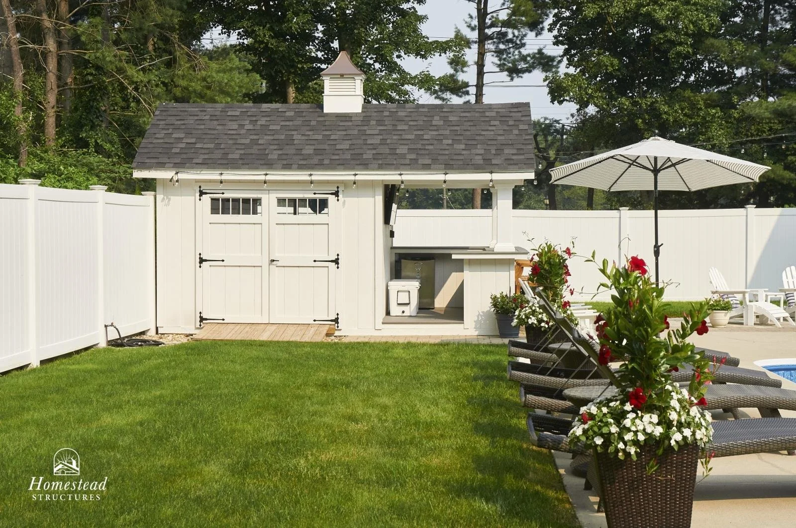 A backyard with a white fence, green lawn, and a small white shed with a gray shingled roof. There are patio chairs, a large white umbrella, and flower pots with red and white flowers.
