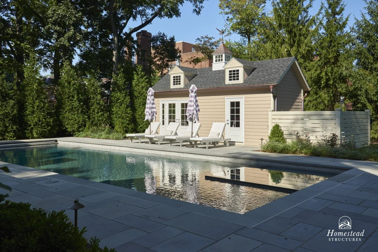 A backyard pool area with a small beige poolhouse, four lounge chairs with striped umbrellas, surrounded by greenery and trees, with a fence on one side and a large house visible in the background.