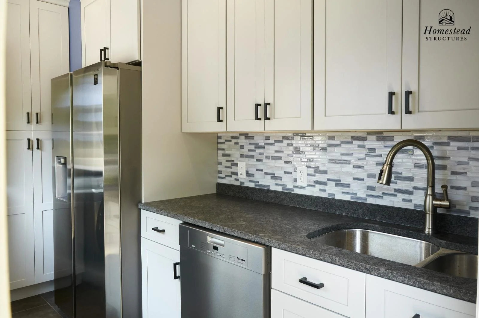 Modern kitchen with white cabinets, a stainless steel refrigerator, a granite countertop, a mosaic tile backsplash, a built-in dishwasher, and a brass kitchen sink faucet.