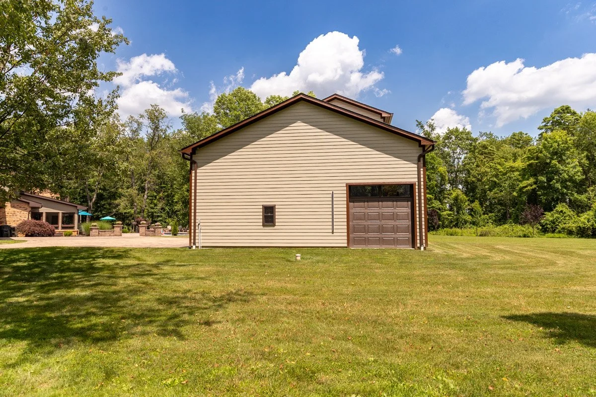 View of a beige building with a brown garage door, small window, and gabled roof, surrounded by green grass and trees, under a partly cloudy sky.