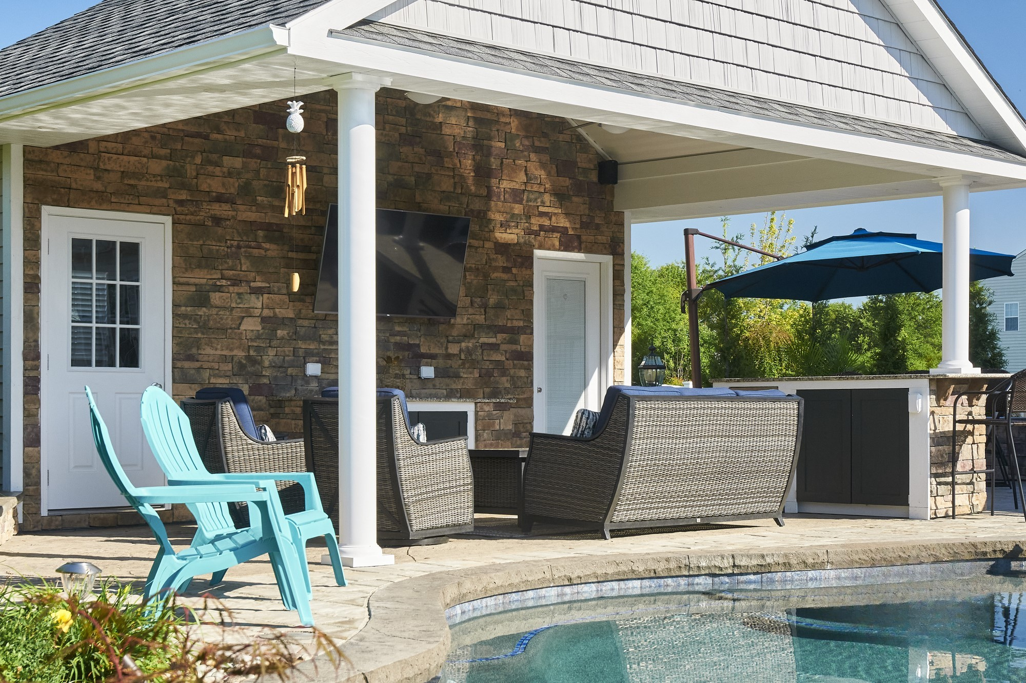 Backyard patio with outdoor seating, a floating TV, a swimming pool, a large blue umbrella, and a house with brick and siding. Green trees in the background.