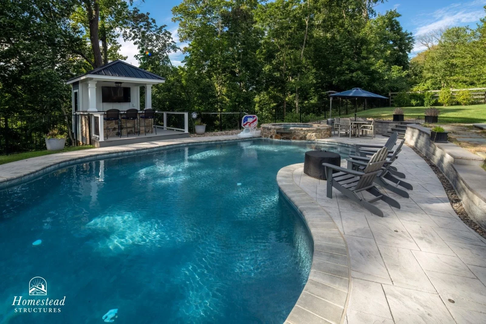 A backyard pool area with a curved swimming pool, a hot tub, and a small covered structure with a television, surrounded by trees and greenery.