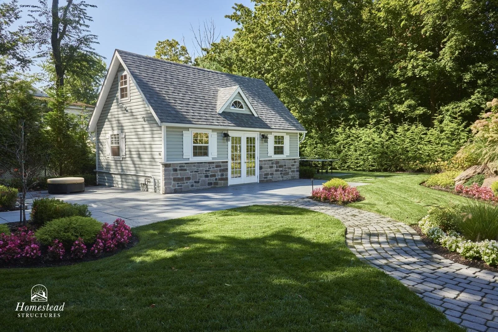 A small house with a stone and wood exterior, surrounded by a landscaped yard with green grass, pink and white flowers, and a curved stone pathway leading to the front of the house. Trees are in the background.