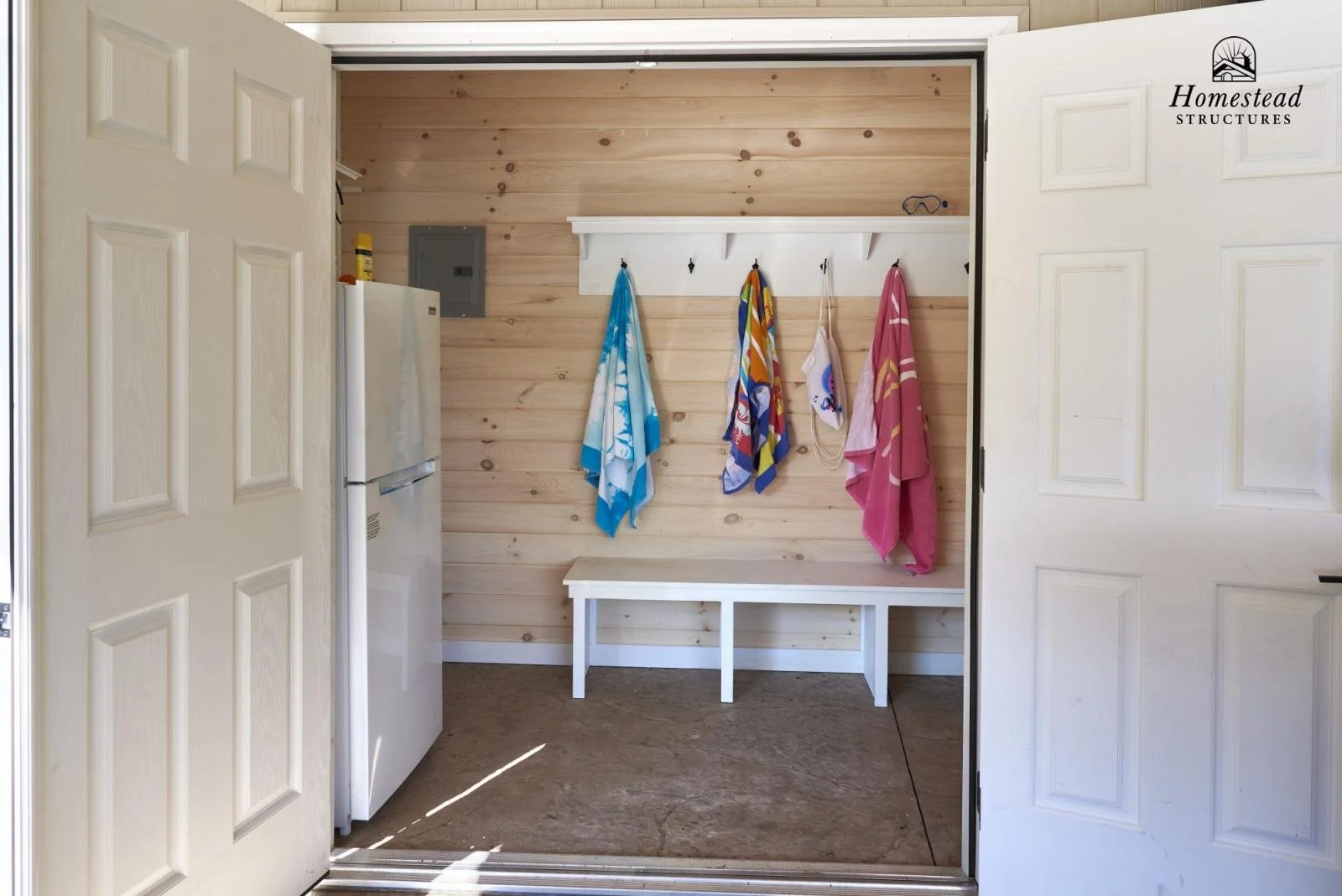View of a small laundry or changing area with a wooden wall, a white bench, colorful towels hanging on hooks, and a small refrigerator.