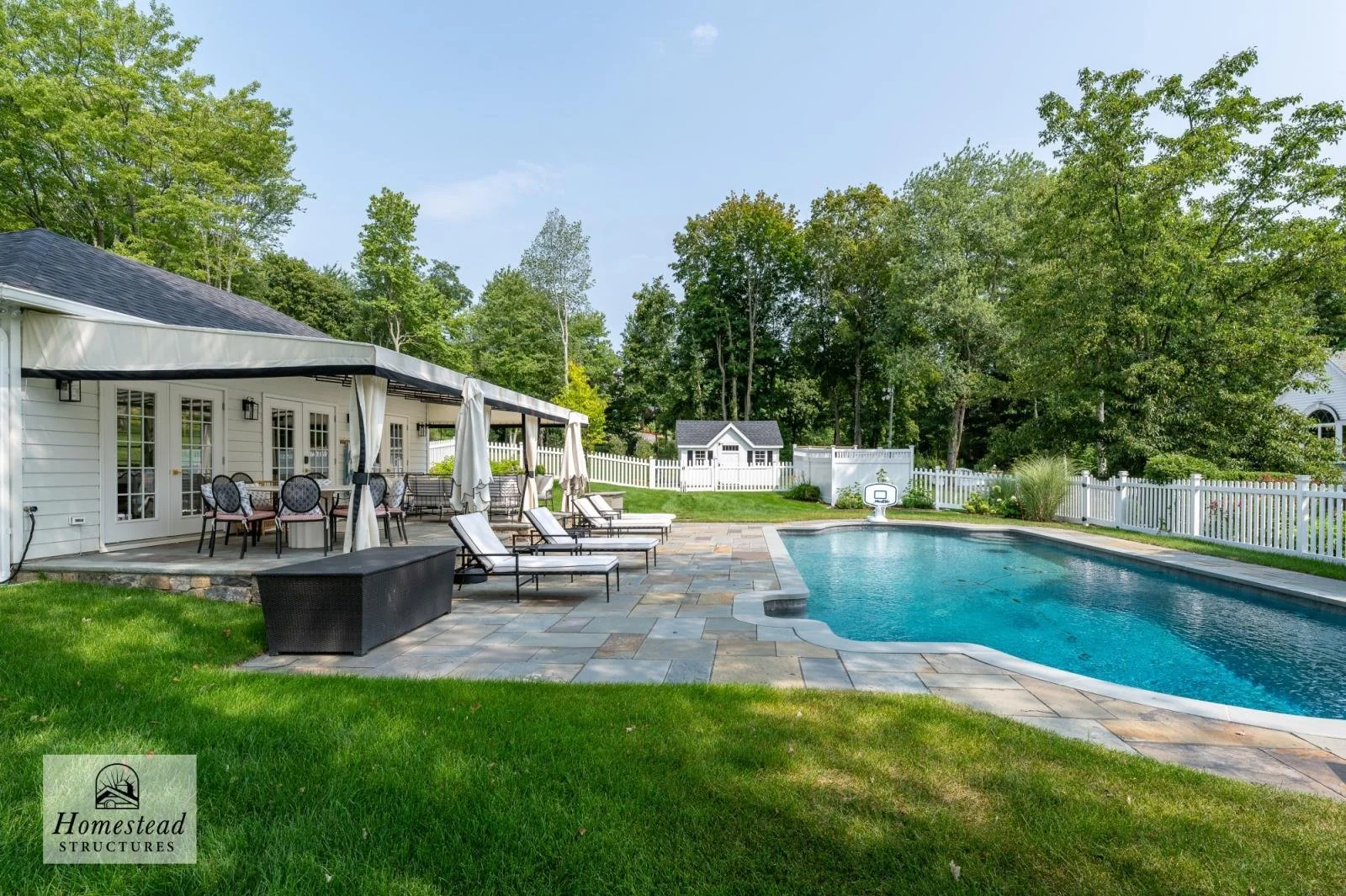 Backyard with a swimming pool, poolside lounge chairs, a covered patio area with outdoor dining table and chairs, surrounded by a white picket fence, lush green grass and tall trees in the background.