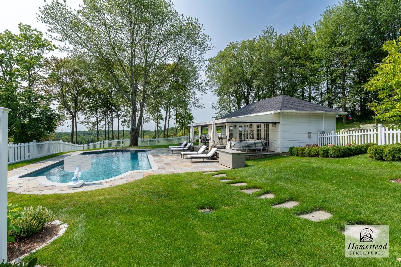 Backyard with swimming pool, patio, lounge chairs, and a gazebo, surrounded by green grass, trees, and a white fence.