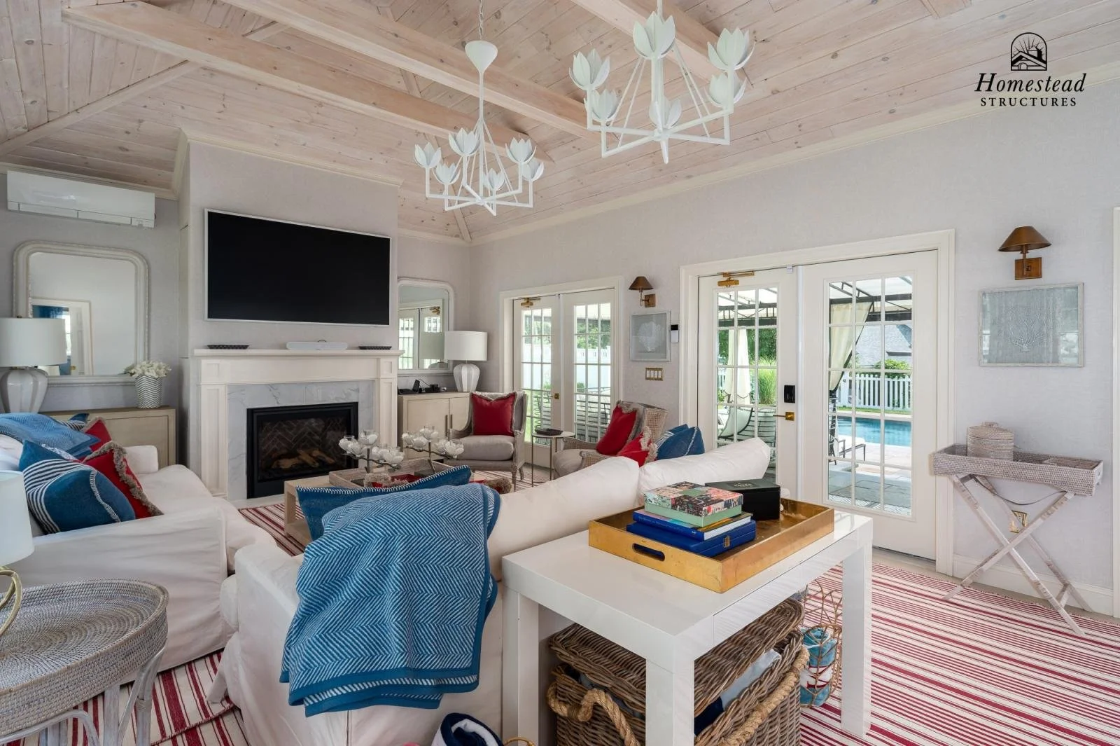 Living room with white walls, wood-paneled ceiling, and large windows leading to patio and pool. Contains white sofas with blue and red pillows, coffee table, side tables, lamps, fireplace, and a mounted TV. Decor includes mirrors, artwork, and a cha