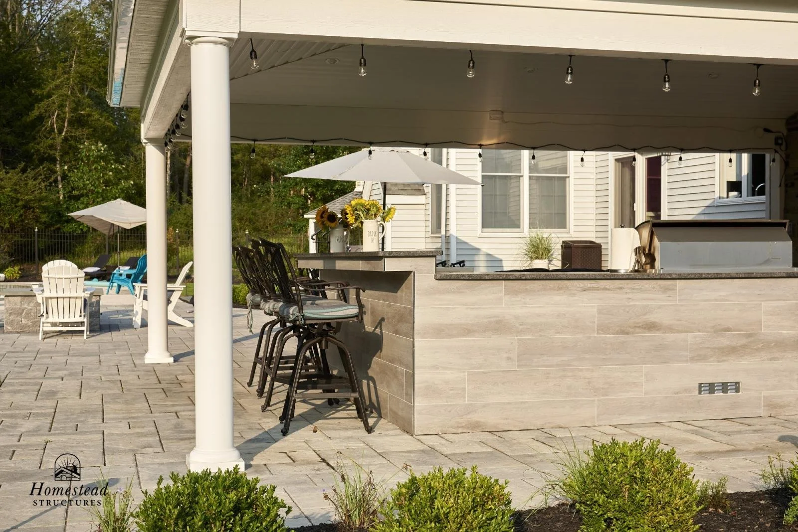 Outdoor patio area with bar counter, bar stools, white Adirondack chairs, umbrellas, and decorative sunflower vases.