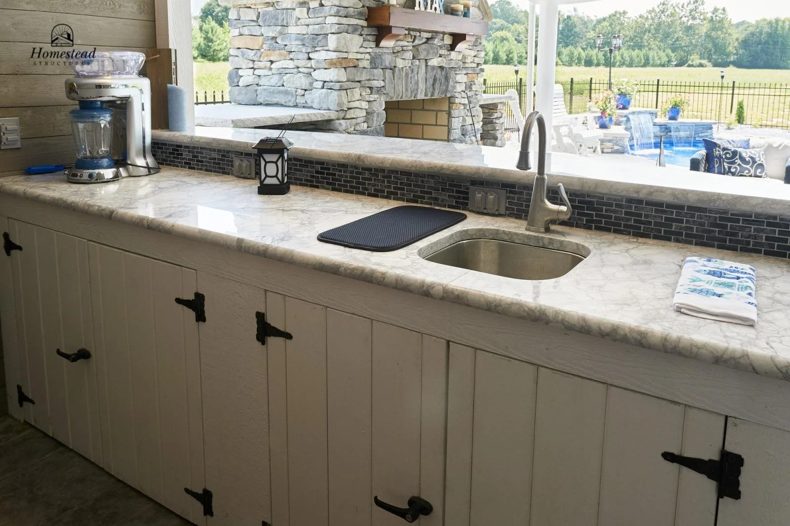 Kitchen counter with a coffee maker, a black mat, a towel, and a sink, overlooking a patio with a pool and outdoor furniture outside.