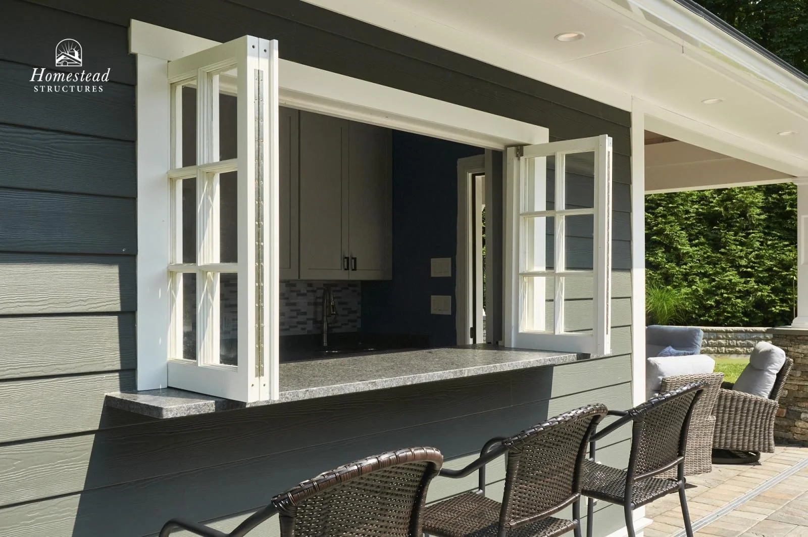Outdoor kitchen with open window, gray cabinets, black countertop, and patio furniture with chairs. Green trees and stone wall in background.