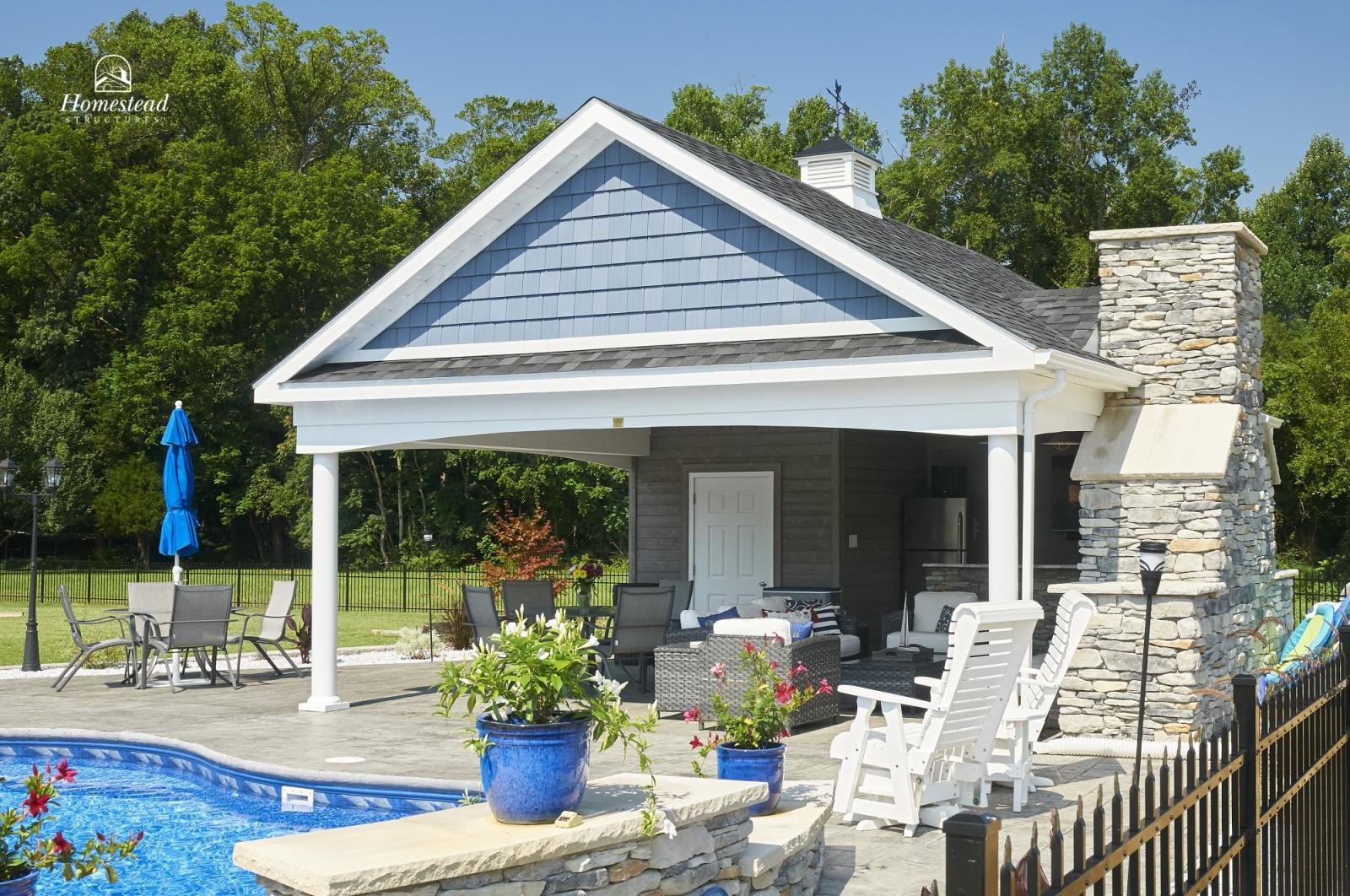A backyard with a pool, outdoor furniture, and a covered patio area featuring a stone chimney and a blue and white house structure.