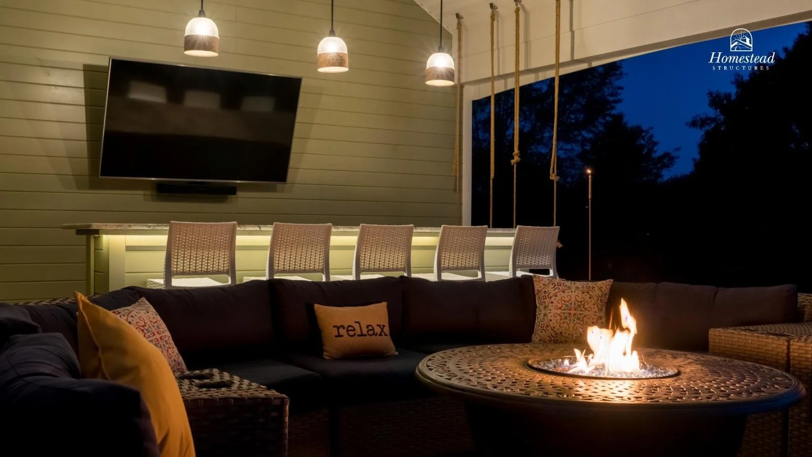 An outdoor seating area with a lit firepit, a dark outdoor sectional sofa with decorative pillows, a wall-mounted television, and pendant lights overhead. The scene is set during nighttime with a dark sky and trees in the background. The image includ
