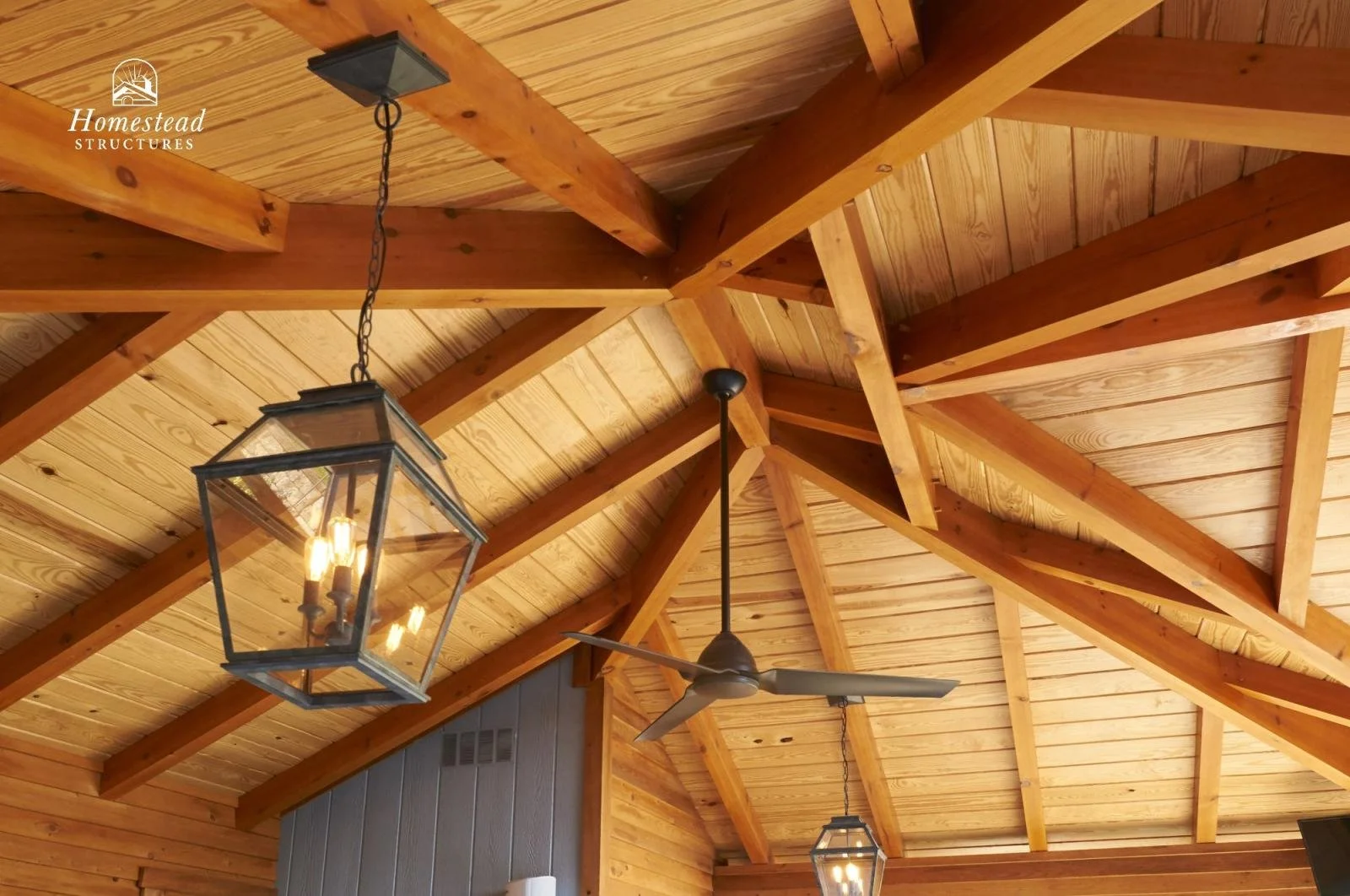 Interior view of a wooden ceiling with exposed beams, two hanging lantern-style light fixtures with candles, and a ceiling fan, part of a homestead-style room.